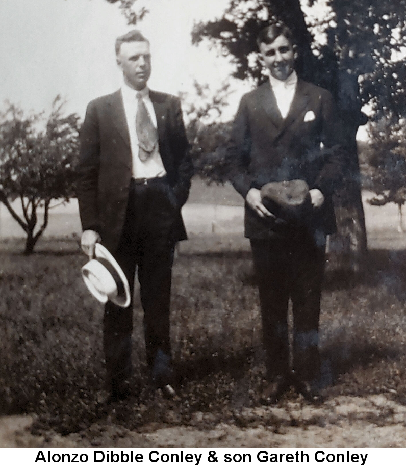 Black and white photo of Alonzo Dibble Conley, standing in front of trees in a black suit with open jacket showing a white shirt and short wide tie, holding a white fedora at his knee, next to his son Gareth Conley, in black suit and white bow tie, holding a dark object with both hands at his waist.